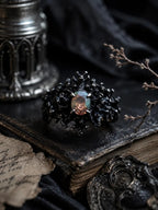 Dark Bloom Ring — black oxidized 925 sterling silver bloom of thorns and coral around an iridescent oval opal, on a dark book